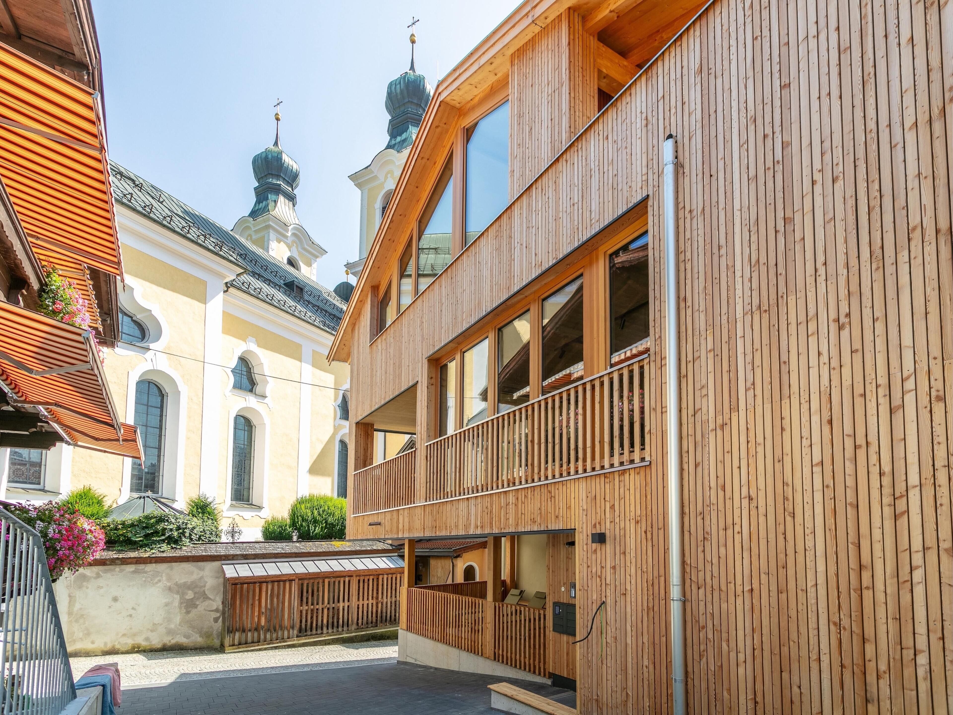 Ein hölzernes Gebäude mit Balkon, großen Fenstern und einem traditionellen Bauwerk im Hintergrund.