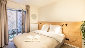 Hotellihuone, jossa on sänky, puinen pääty, yöpöytä, peili ja lasiovesta avautuva näkymä ulos.
