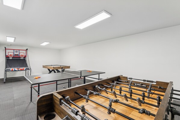 A room with a foosball table, ping pong table, and a basketball hoop.