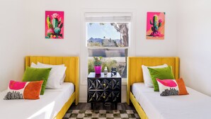 Une chambre avec deux lits, chacun doté d’une tête de lit jaune, de draps blancs et d’oreillers colorés. Les murs sont ornés de deux tableaux représentant des cactus. Une fenêtre offre une vue sur les montagnes, et une petite table est agrémentée d’un pot violet et d’une table d’appoint noire aux motifs géométriques.