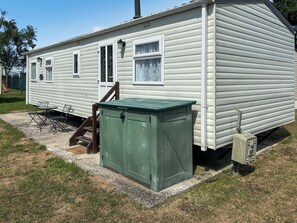Une maison mobile de plain-pied avec un abri de jardin vert, un petit patio doté de meubles d’extérieur et un escalier apparent menant à l’entrée.