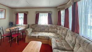 A living room with a curved sofa, a wooden table, and a dining area with chairs. - 2 bedroom accommodation in Burnham-on-Sea (Burnham-on-Sea)