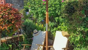 Deux fauteuils de jardin en bois, dotés de coussins blancs et surmontés d’un chapeau, sont placés dans un jardin où l’on trouve diverses plantes et, au fond, un mur de pierres.