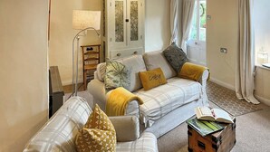 Un salon comprenant un canapé, une table d’appoint, une lampe, un meuble et une fenêtre avec des rideaux.