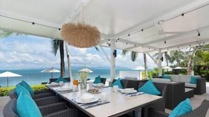An outdoor dining area with a view of the ocean, white tablecloths, and blue cushions.