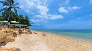 A beach with large rocks, a palm tree, and a building with umbrellas.