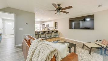 Un salon moderne avec un canapé en cuir marron, un plaid blanc et une table basse. On y trouve également un ventilateur de plafond et un téléviseur à écran plat fixé au mur.