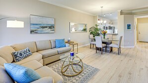 Een woonkamer met een beige bank, een glazen salontafel en een eethoek met een tafel en stoelen.