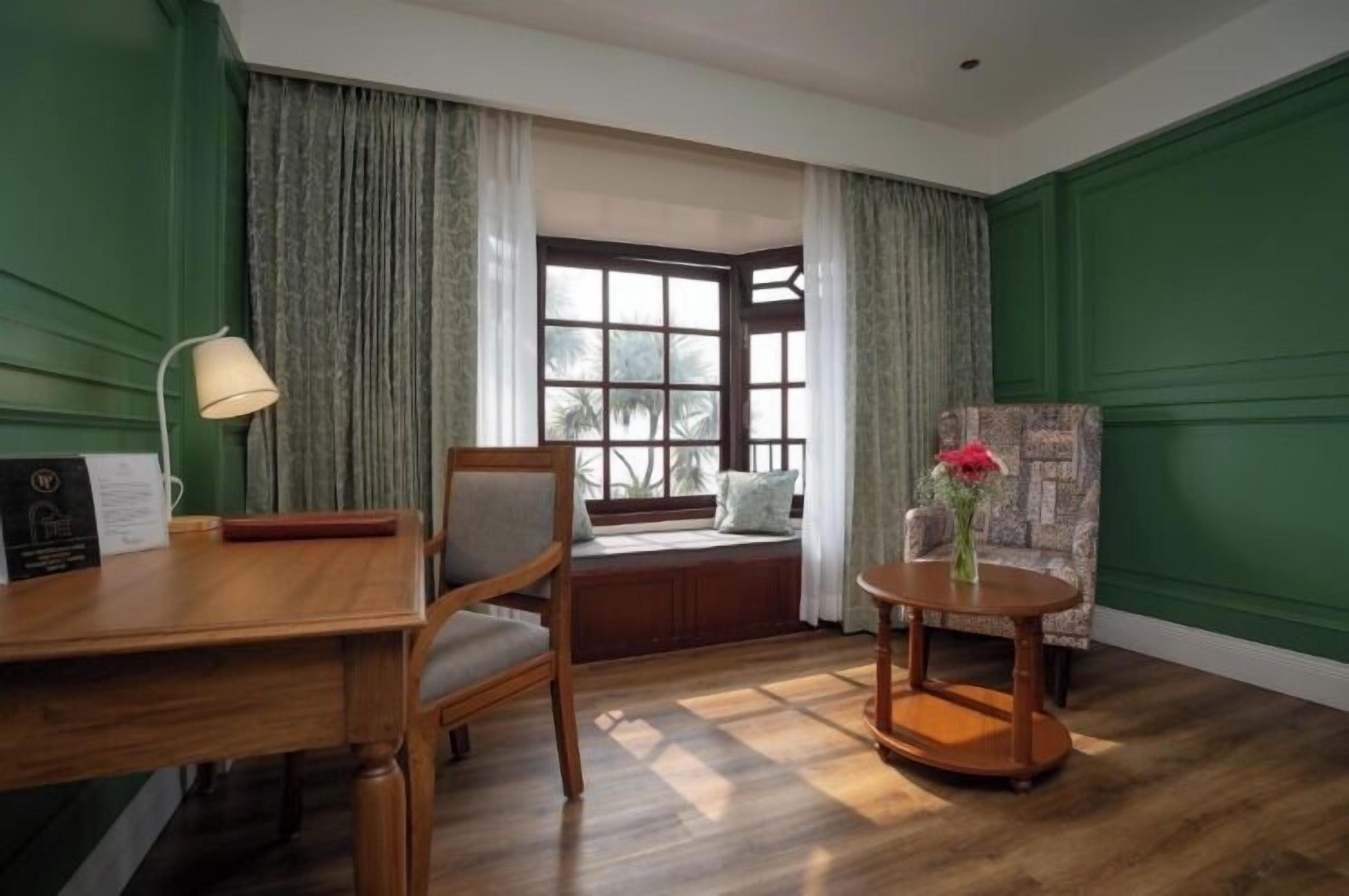 Een kamer met een houten bureau, een stoel, een ronde tafel met een vaas en een raam met gordijnen.