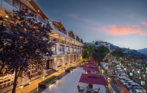 Un hotel con estructura de varios pisos, mesas al aire libre con sombrillas y vistas de la ciudad circundante.