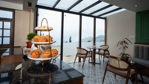 Montaje para el desayuno con un expositor escalonado que contiene muffins y pasteles, una máquina de café y una planta en maceta. Se observan sillas y una mesa en primer plano, y una ventana grande con vistas al exterior.