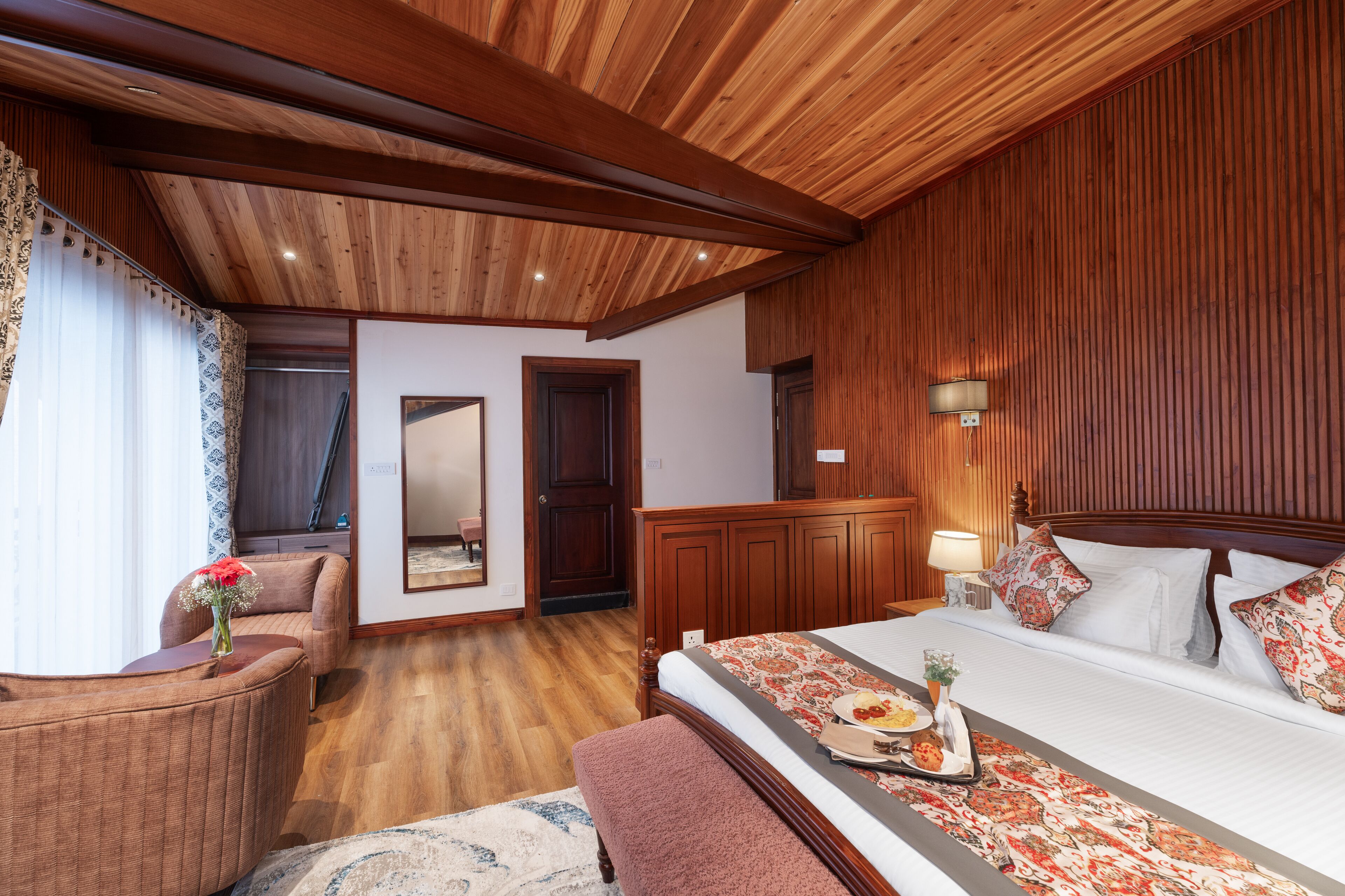 A hotel room with a wooden ceiling, a large bed with a floral bedspread, a sitting area with a chair and a vase of flowers, and a wooden wardrobe.