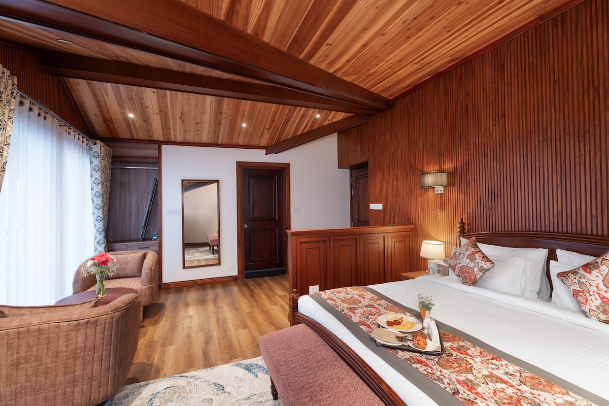 A hotel room with a wooden ceiling, a large bed with a floral bedspread, a sitting area with a chair and a vase of flowers, and a wooden wardrobe.