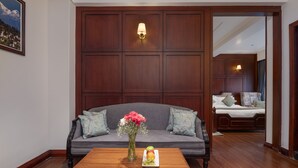 Una habitación con pared de paneles de madera, un sofá gris con cojines florales, una mesa de centro de madera con un jarrón de flores y fruta, y una cama al fondo.