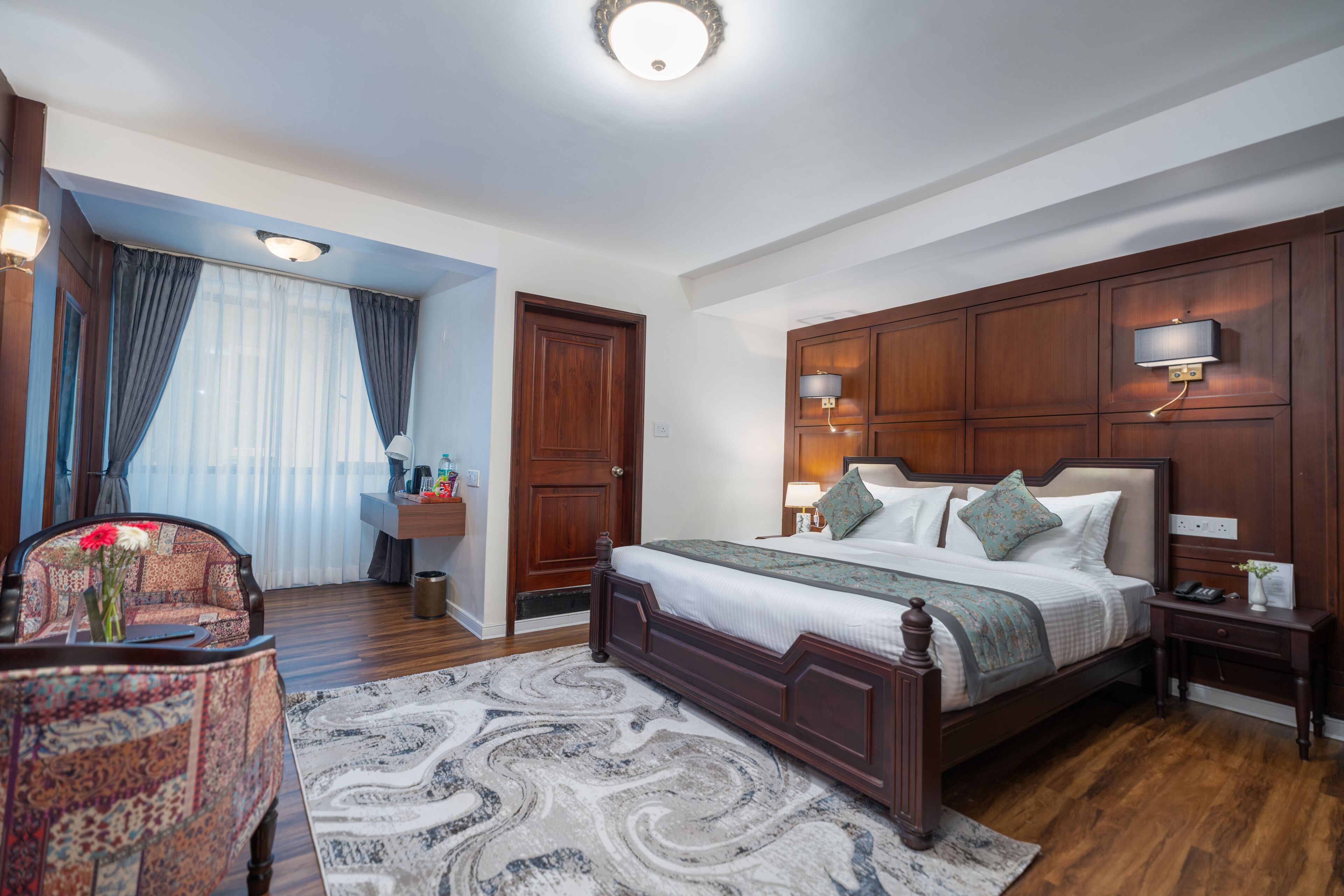 A bedroom with a large bed, wooden headboard, bedside lamps, a nightstand, a desk, and a chair.