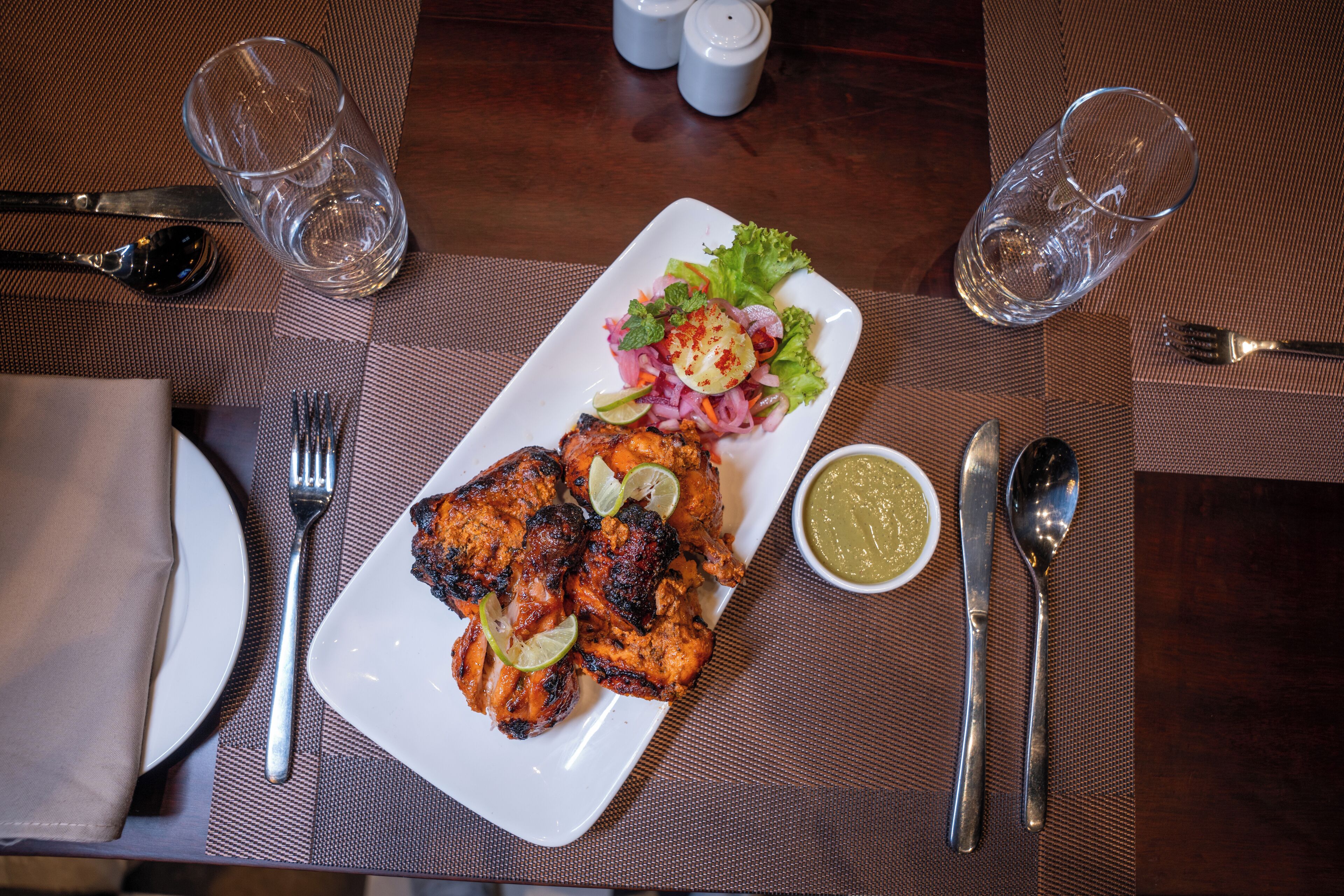 A dining table with a plate of grilled chicken, a salad, and a small bowl of sauce.
