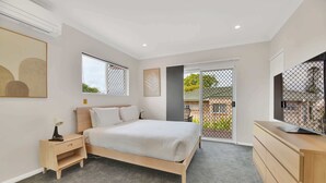 A bedroom with a bed, bedside table, lamp, dresser, TV, and a view of the outside. - Tranquil 3-Bed Unit with Lush Garden Views (Taringa)