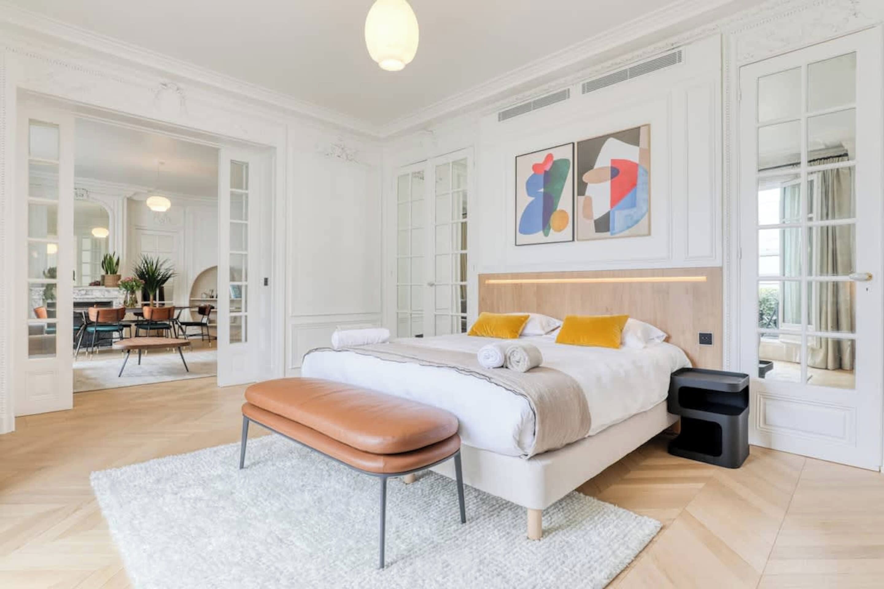 A spacious bedroom with a large bed, a bench, and a view of the dining area.