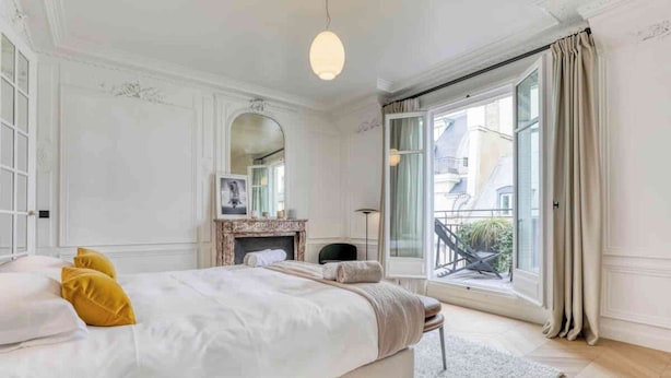 A bedroom with a large bed, a fireplace, a balcony with a view, and a large window with curtains.
