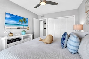 Un dormitorio con cama, televisor mostrando una escena de playa, ventilador de techo y puerta que da a otra habitación.