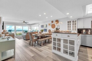 A spacious living area with a large window overlooking the ocean, a dining area with a wooden table and chairs, and a kitchen with white cabinetry and a built-in island.