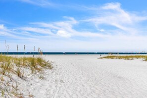 Een strand met wit zand en weinig gras, een helderblauwe lucht en de oceaan op de achtergrond.