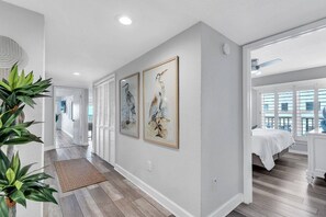 A hallway with a wooden floor, a white door, and a bed with a white bedspread in the adjacent room.