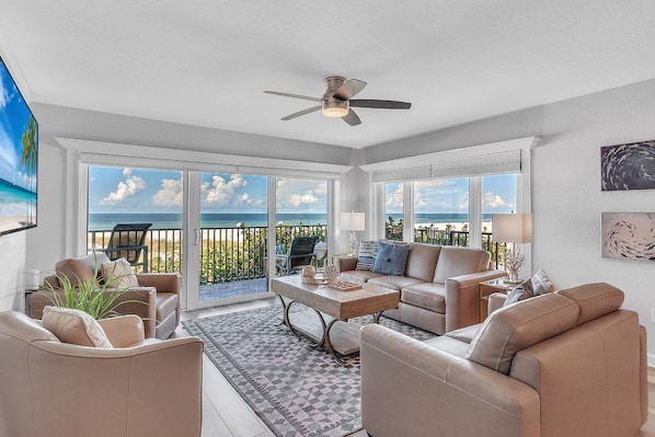 A spacious living room with a large window overlooking a beach, a ceiling fan, and a view of the ocean.