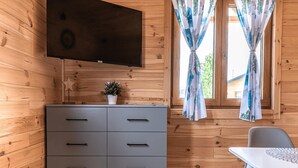 Un intérieur en bois avec un meuble gris, un téléviseur à écran plat, une petite plante et une fenêtre avec des rideaux.