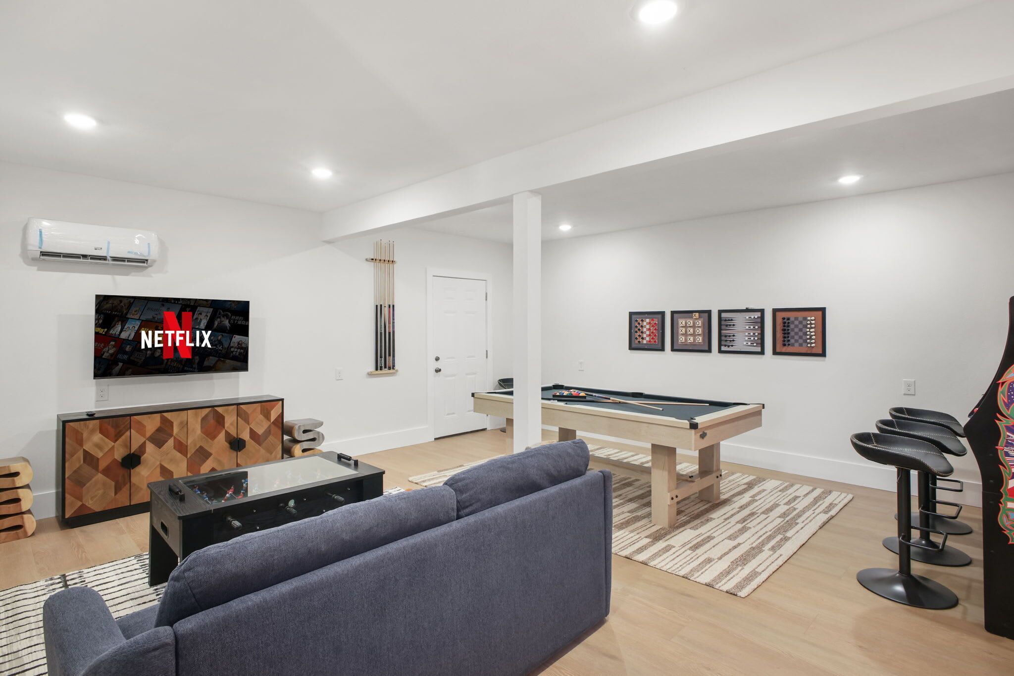 A modern living room with a pool table, a sofa, a bar area, and a mounted TV displaying the Netflix logo.
