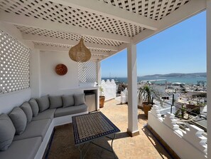 Een balkon met een witte bank, een tafel met patroon en uitzicht op zee en stadsbeeld.