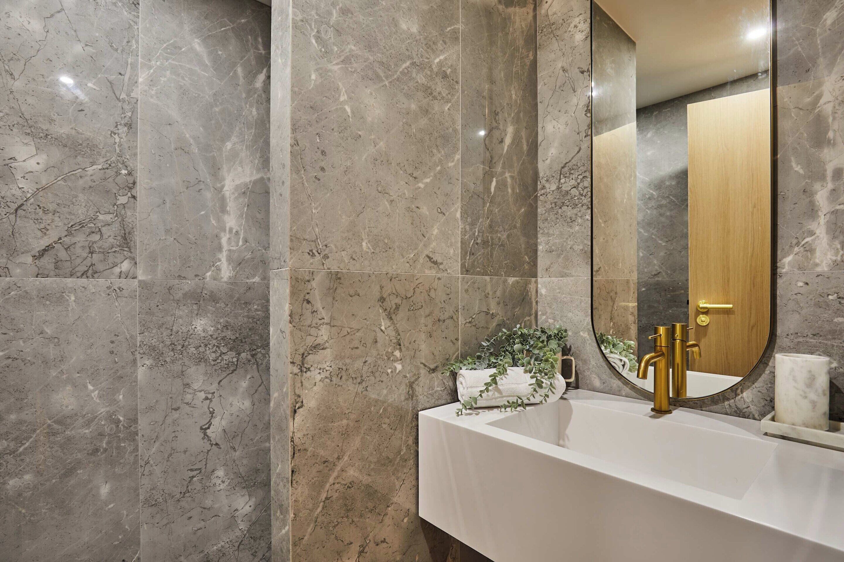 A bathroom with a marble wall, a white sink, a gold faucet, a round mirror, and a wooden door.