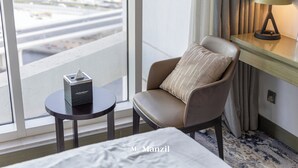 A hotel room with a bed, a chair with a pillow, a small table with a tissue box, a lamp, and a view of a cityscape through a window. - Manzil-Waterfront Studio in Business Bay (Dubai)