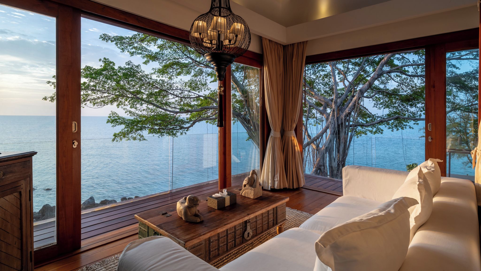 A room with a large window overlooking a body of water, a wooden floor, a white sofa, a wooden coffee table, and a hanging light fixture.