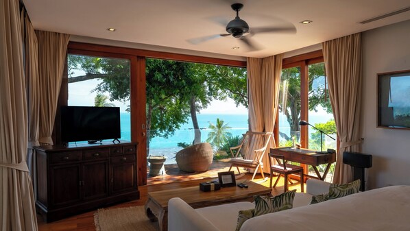 A bedroom with a large bed, a television on a wooden cabinet, a desk with a chair, and a view of the ocean.