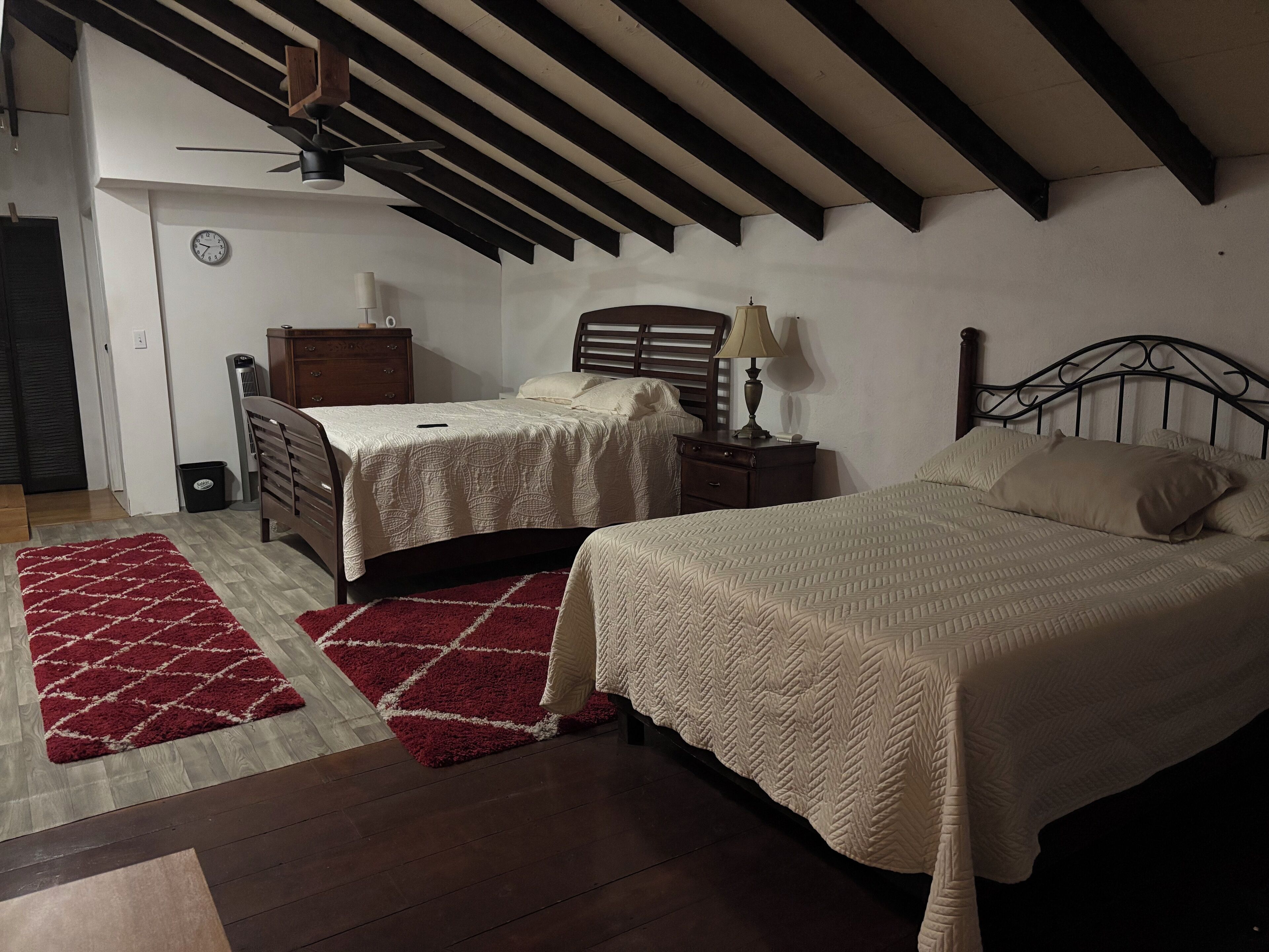A bedroom with two beds, a wooden floor, a red rug, a ceiling fan, and a clock on the wall.