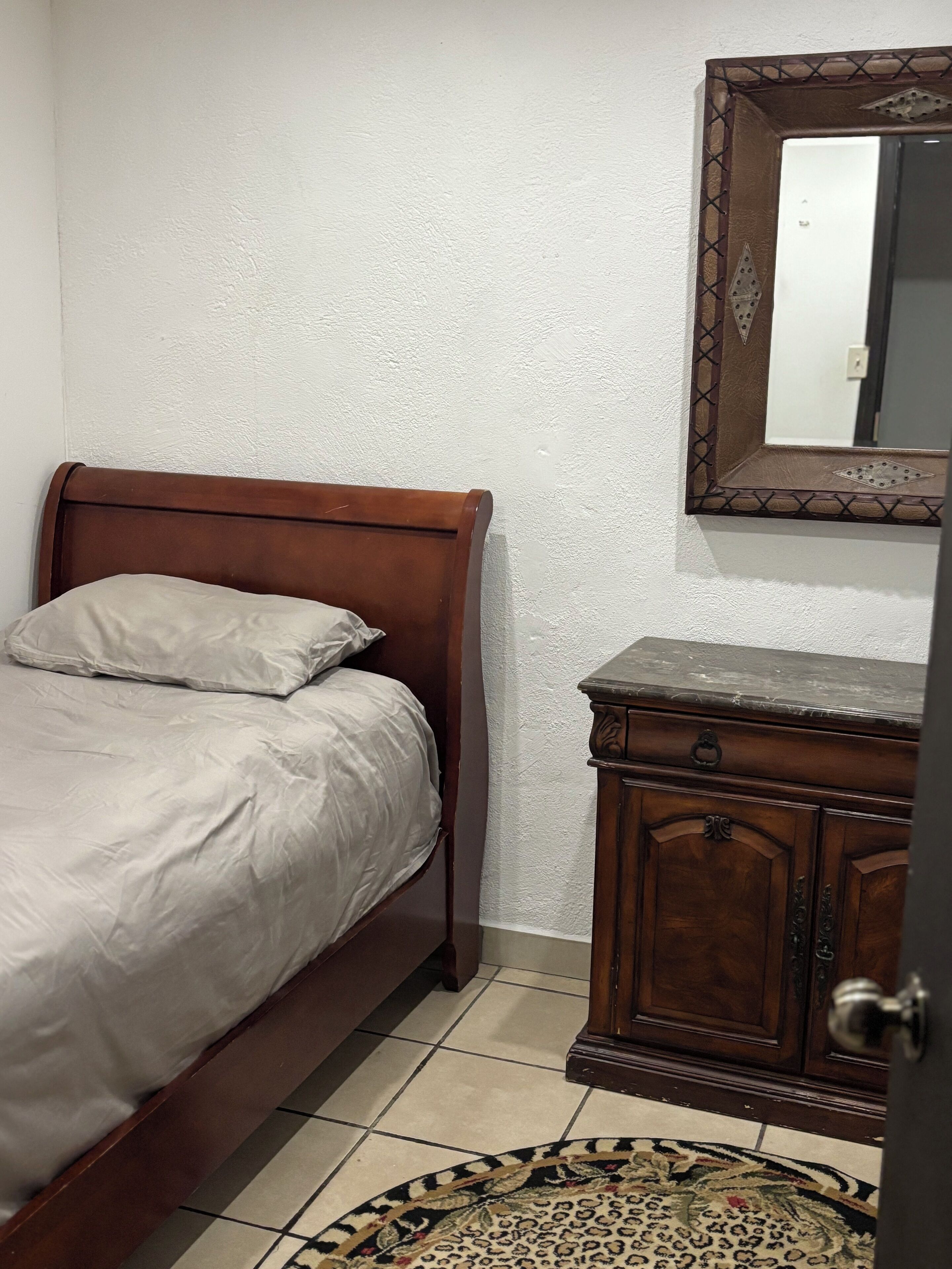 A bedroom with a wooden bed, a small wooden dresser, and a mirror on the wall.