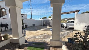 A covered patio area with a stone floor, potted plants, a barbecue grill, and a white vehicle parked outside. - Casa Vacacional Marqués (Playas de Rosarito)