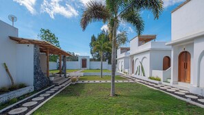 A courtyard with a palm tree, stone pathways, and white buildings with wooden doors. - Pulestis Glamping (Kintamani)