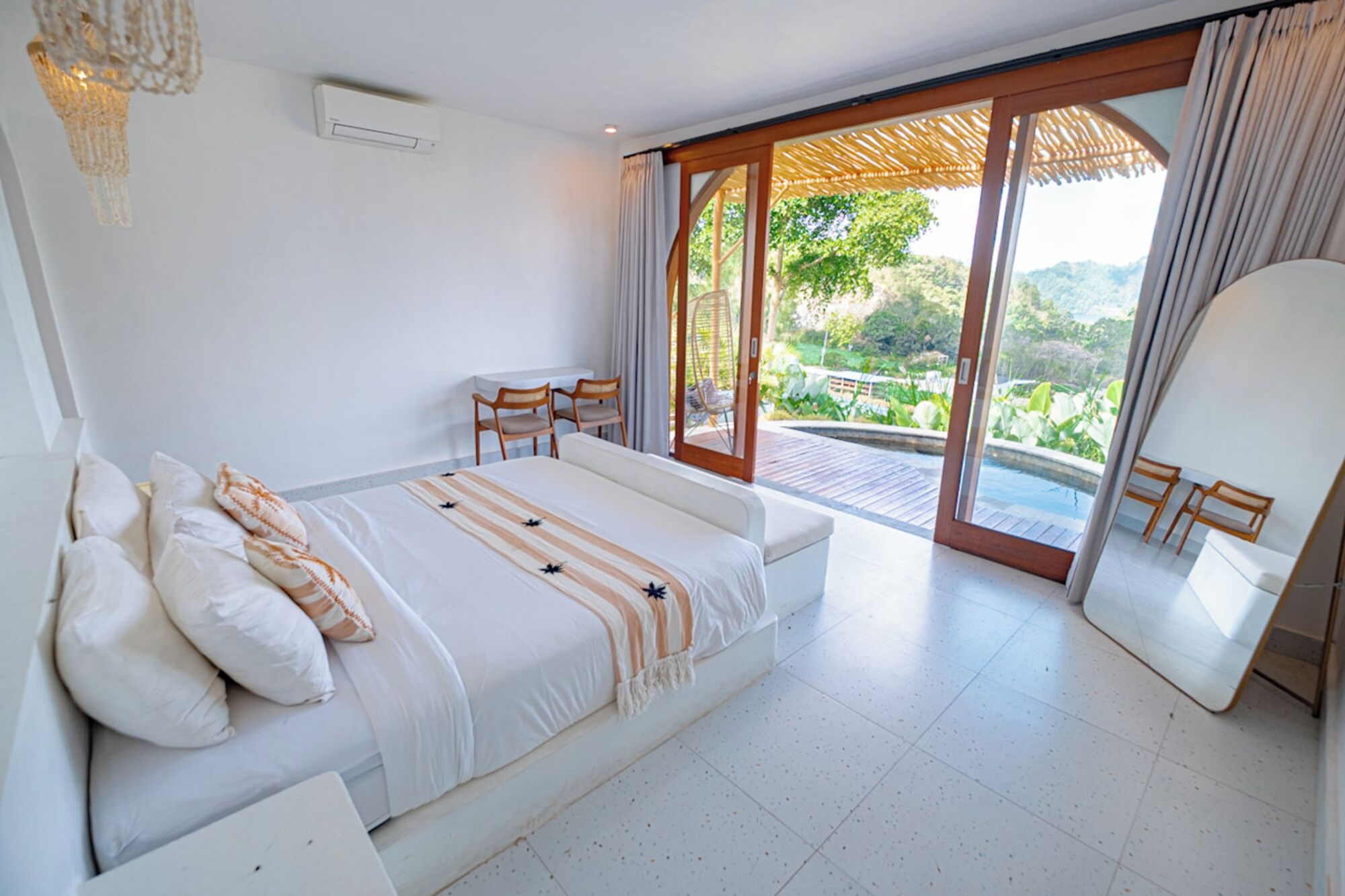 A bedroom with a bed, a desk, and a chair, a sliding glass door leading to a balcony with a view of greenery.