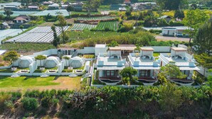 An aerial view of a resort with multiple buildings, including a large central structure with a flat roof and smaller buildings with sloped roofs. There are well-maintained gardens and fields surrounding the buildings. - Pulestis Glamping (Kintamani)