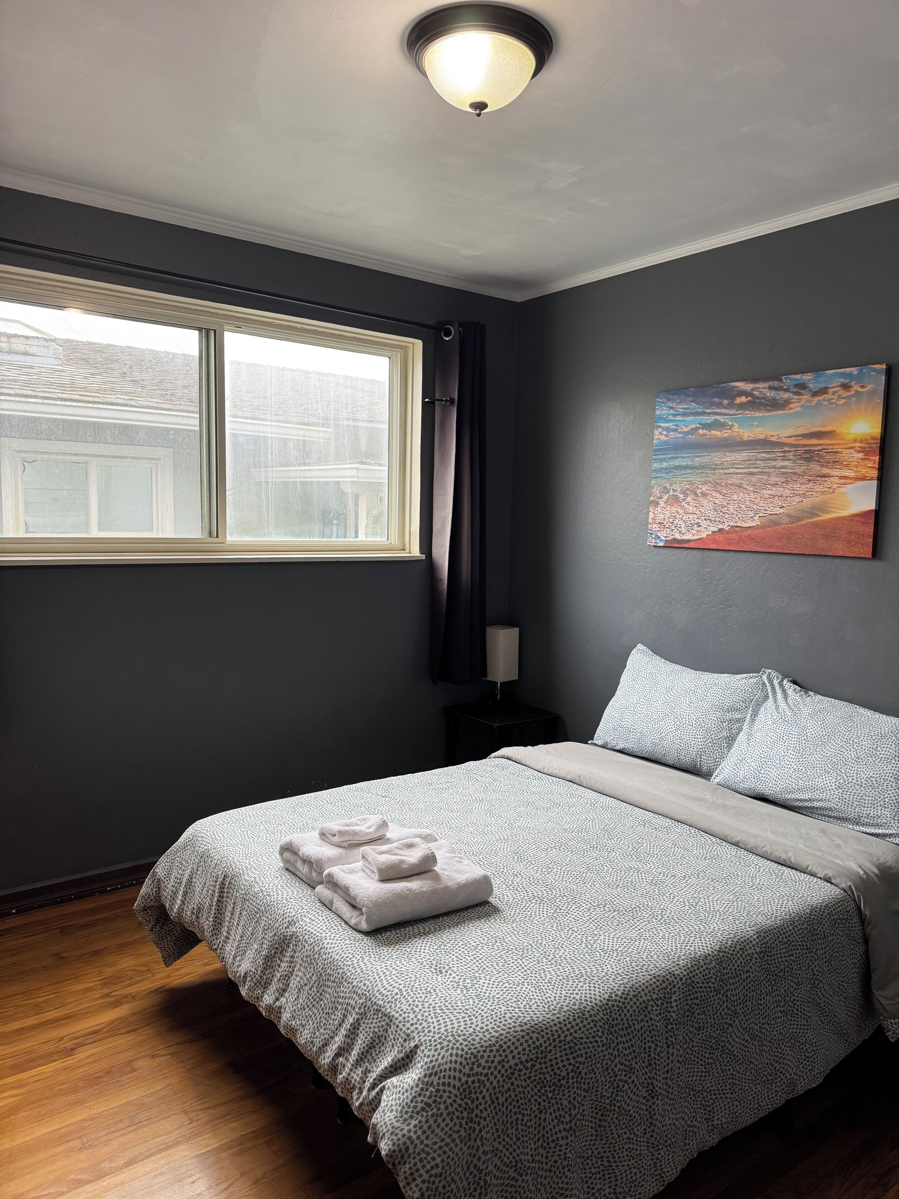 A bedroom with a large window, a bed with a patterned bedspread, a painting of a sunset, and a bedside lamp.