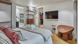 A modern bedroom with a large bed, a wooden nightstand, a wall-mounted TV, and a unique brown chair. - Charming Apartment Faubourg St Honore Madeleine (Paris)