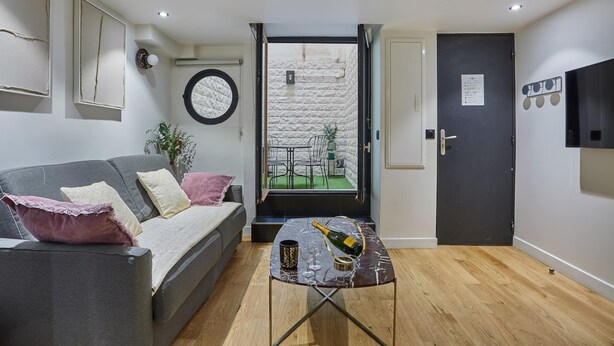 A compact living space with a sofa, a small table with a bottle and glass, a door leading to an outdoor area, and a wall-mounted TV. - Charming Apartment Faubourg St Honore Madeleine (Paris)