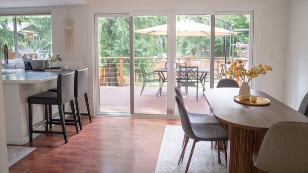 A modern kitchen with a bar area, a dining table with chairs, and a sliding glass door leading to an outdoor patio with umbrellas and chairs. - Forest Escape – Hiking & Rattlesnake Lake Nearby (North Bend)
