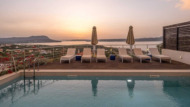 A rooftop pool with lounge chairs and umbrellas overlooking a scenic view of a body of water and distant mountains during sunset.