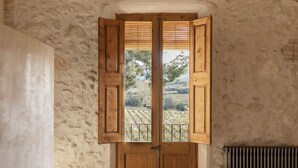 A bedroom with a stone wall, wooden beams, a large window with wooden shutters, a radiator, and a hanging light fixture. - Boutique Manor Farmhouse, pool & vineyard views, 35' Barcelona, 25' Sitges (Sant Pau d'Ordal)