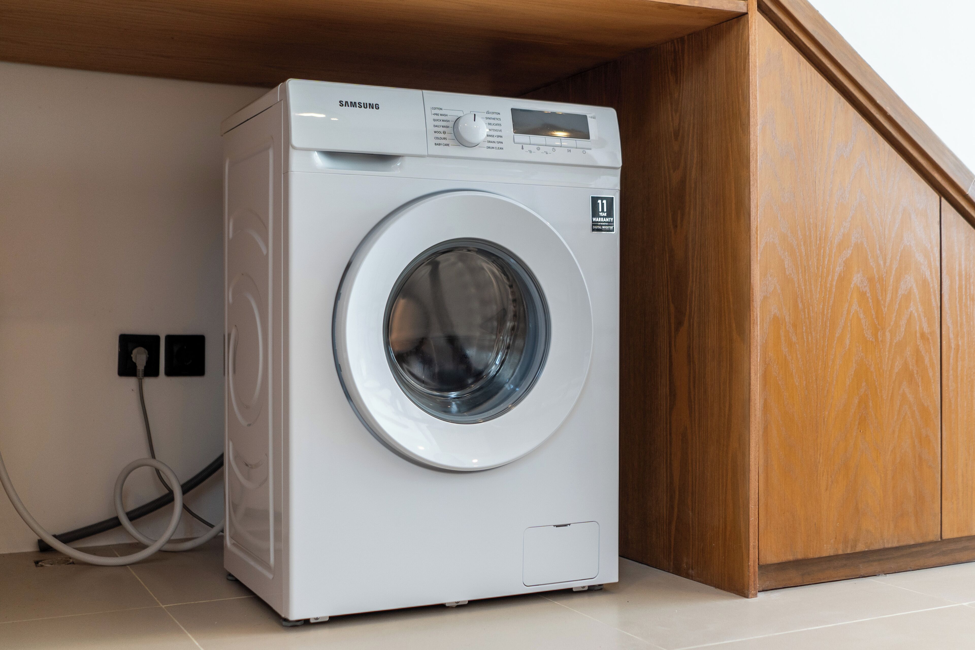 A white Samsung washing machine in a room with a wooden cabinet and a tiled floor.