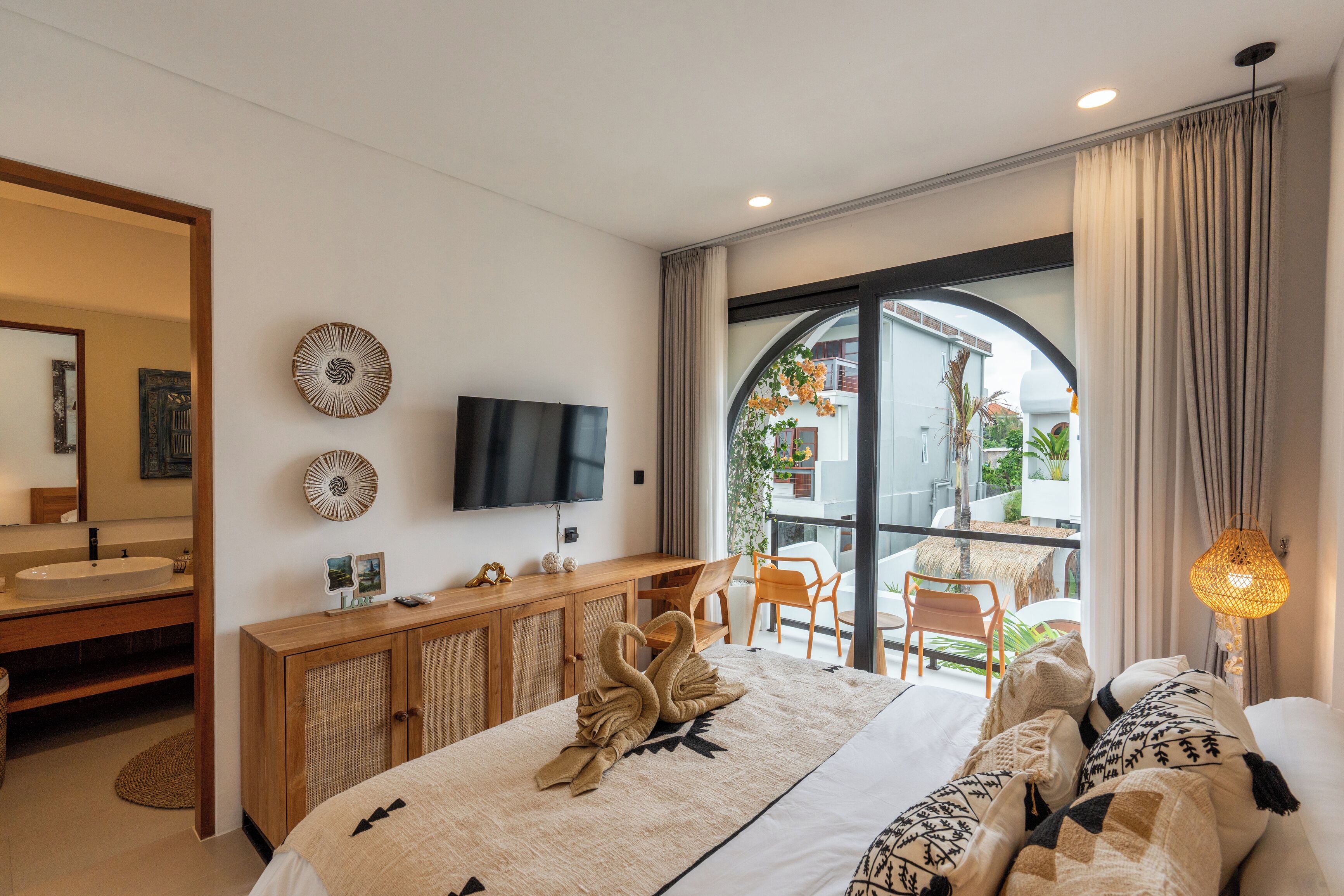 Habitación de hotel moderna con una cama grande, una mesita de madera, televisor y balcón con mobiliario exterior.