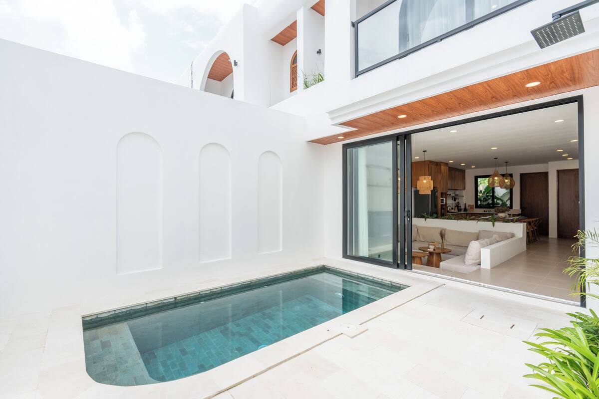 Ett modernt hus med en swimmingpool, en glasdörr som leder in till ett vardagsrum och ett kök som syns genom glaset.